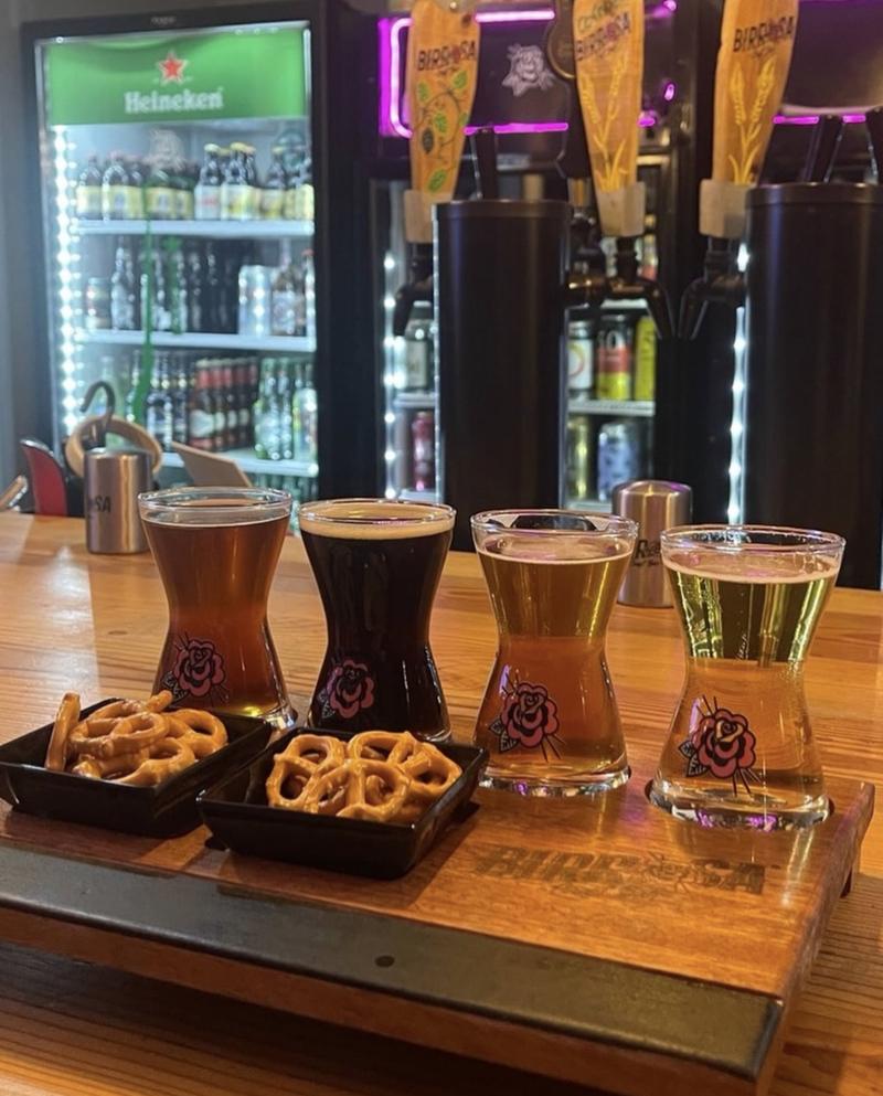 Flight de cuatro vasos de cerveza sobre una tabla de madera con pretzels, frente a los grifos de Birrosa